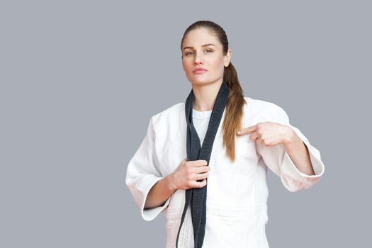 Proud attractive athletic woman in white kimono standing and wraps black belt around the neck and pointing finger. Japanese karate and sports concept. indoor studio shot, isolated, grey background
