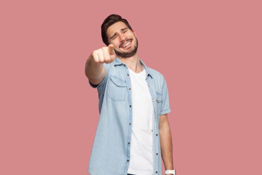 Portrait of funny happy handsome bearded young man in blue casual style shirt standing pointing and looking at camera with toothy smile. indoor studio shot, isolated on pink background.
