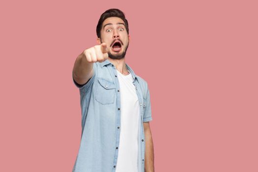 Portrait of shocked handsome bearded young man in blue casual style shirt standing pointing and looking at camera with surprised unbelievable face. indoor studio shot, isolated on pink background.