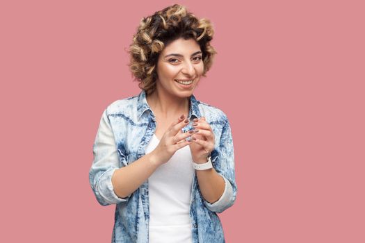Portrait of funny cunning young woman with curly hairstyle in casual blue shirt standing and looking at camera with toothy smile playful face. indoor studio shot, isolated on pink background.