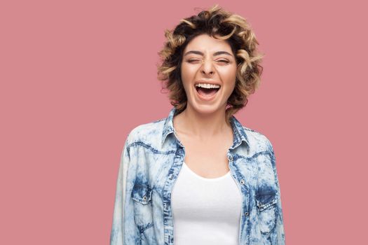Portrait of happy laughter young woman with curly hairstyle in casual blue shirt standing, clossed eyes and looking at camera and laughing and enjoy. indoor studio shot, isolated on pink background.