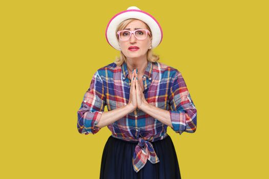 Please forgive me. or give me one more chance, Portrait of hopeful mature woman in casual style with hat and eyeglasses standing with palm hands and looking at camera, begging and asking. studio shot.