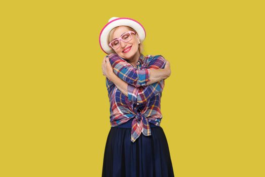 Portrait of peacful happy modern stylish mature woman in casual style with hat, eyeglasses standing, hugging herself with closed eyes and toothy smile. indoor studio shot isolated on yellow background