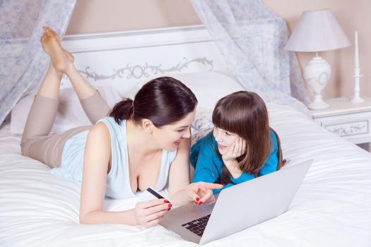 Happy mother and daughter do online shopping and pay with credit card with laptop lying on bed in bedroom. Happy family. Studio shot.