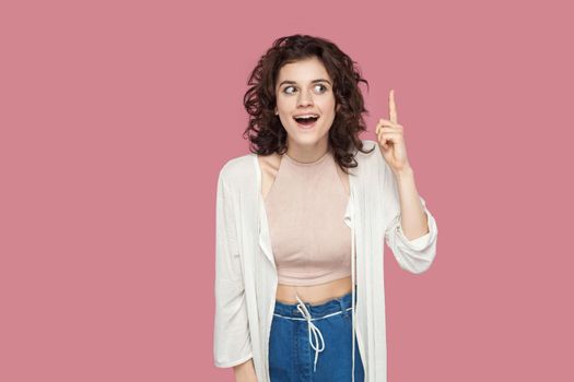 Idea. Portrait of surprised beautiful brunette young woman with curly hairstyle in casual style standing, holding finger up and excited with new idea. indoor studio shot isolated on pink background.