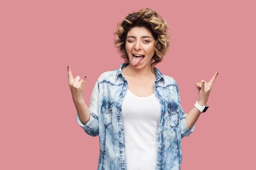 Portrait of happy funny young woman with curly hairstyle in casual blue shirt standing, winking, tongue out and looking at camera with rock horns sign. indoor studio shot, isolated on pink background.