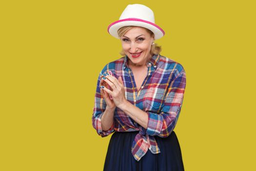 Portrait of cunning modern stylish mature woman in casual style with hat and eyeglasses standing in cunning gesture and looking at camera with smile. indoor studio shot isolated on yellow background.