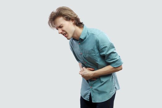 Stomach pain. profile side view portrait of handsome long haired blonde young man in blue casual shirt standing touching his painful belly. indoor studio shot, isolated on light grey background.