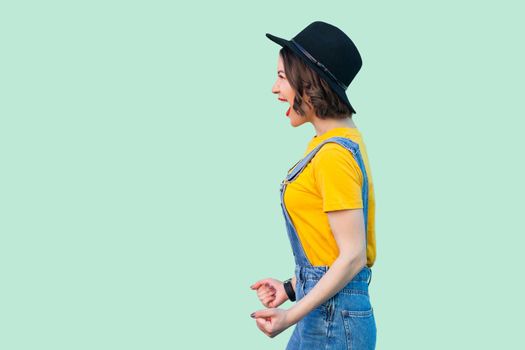 Profile side view portrait of surprised pretty young hipster girl in blue denim overalls, yellow shirt and black hat standing and screaming. indoor studio shot isolated on light green background.