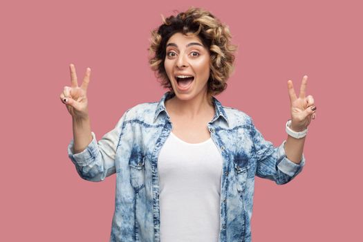 Portrait of happy funny young woman with curly hair in casual shirt standing with victory or peace sign gesture and looking at camera with amazed face. indoor studio shot, isolated on pink background.