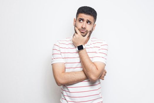 Portrait of pensive handsome bearded young man in striped t-shirt standing, touching his face, looking aside away and thinking about something. indoor studio shot, isolated on white background.