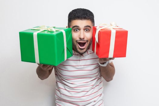 Portrait of amazing shocked attractive young adult man in t-shirt standing, holding two big gift box near face, looking at camera. Studio shot, white background, isolated, indoor