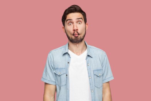 Portrait of funny comic face handsome bearded young man in blue casual style shirt standing with fish lips and looking at camera. indoor studio shot, isolated on pink background.