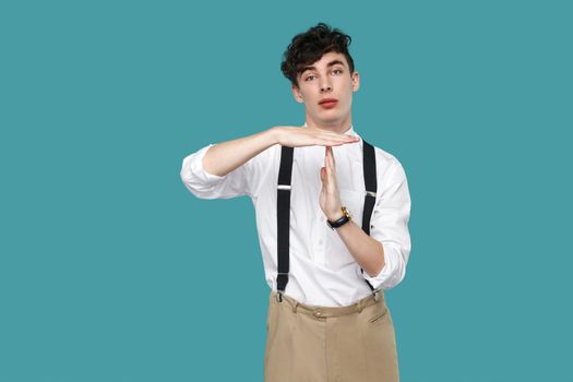 I need more time. man showing timeout gesture. Portrait of handsome hipster curly young businessman in classic casual white shirt and suspender standing. indoor studio shot isolated on blue background