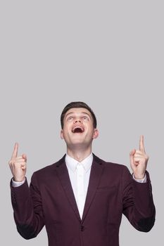 Portrait of surprised handsome young man in violet suit and white shirt, standing, looking top and pointing at copy space on top with amazed face. indoor studio shot, isolated on grey background