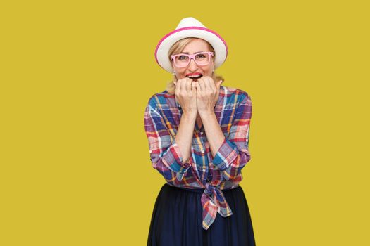 Portrait of sad nervous modern stylish mature woman in casual style with hat and eyeglasses standing, bitting nails and looking at camera and panic. indoor studio shot isolated on yellow background.