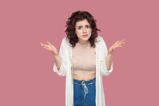 What do you want? Portrait of angry brunette young woman with curly hairstyle in casual style standing, looking at camera with raised arms and asking. indoor studio shot isolated on pink background.