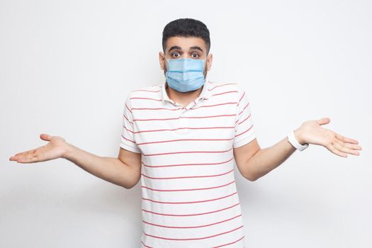 I don't know. Portrait of confused young man with surgical medical mask in striped t-shirt standing with raised arms and dont know what to do. indoor studio shot, isolated on white background.