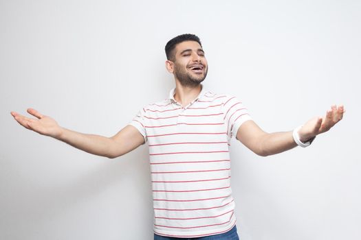Portrait of happy handsome bearded young man in striped t-shirt standing with wide raised arms and looking at camera with toothy smile. indoor studio shot, isolated on white background.