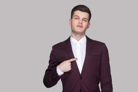 This is me. Portrait of proud handsome young businessman in violet suit and white shirt, standing, looking at camera and pointing himself and bragging. indoor studio shot, isolated on grey background
