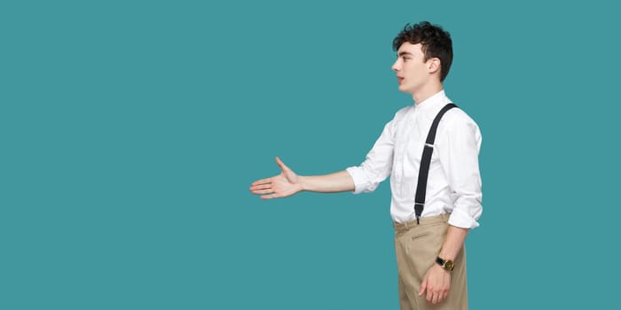 Profile side view portrait of happy handsome curly young businessman in classic casual white shirt standing and giving hand to greeting or handshake. indoor studio shot isolated on blue background.
