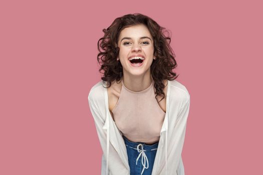 Portrait of excited happy positive beautiful brunette young woman with curly hairstyle in casual style standing, looking at camera and laughing. indoor studio shot isolated on pink background.