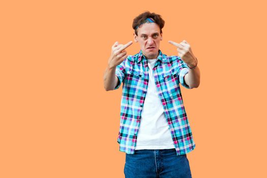Portrait of angry young man in casual blue checkered shirt and headband standing with middle finger fuck sign looking at camera with aggressive face. indoor studio shot, isolated on orange background.