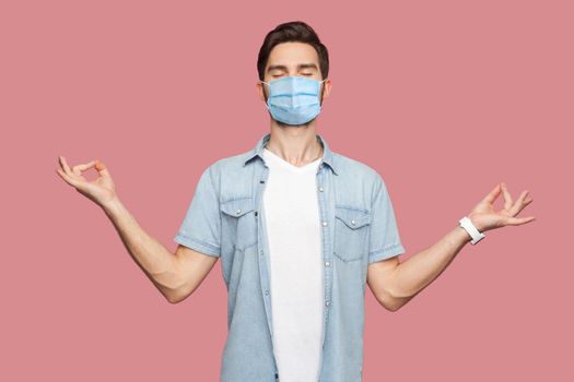 Portrait of calm relaxed young man with surgical medical mask in blue shirt standing with raised arms and doing yoga meditating exercise. indoor studio shot, isolated on pink background.