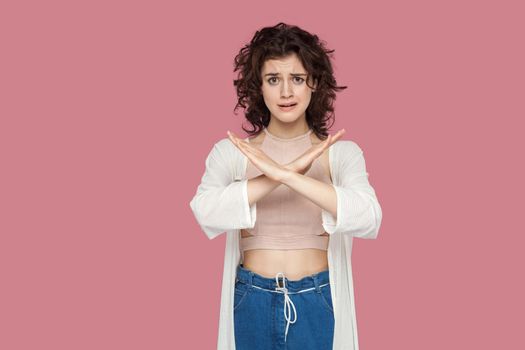 Portrait of dissatisfied beautiful brunette young woman with curly hairstyle in casual style standing and looking at camera wih x sign hands gesture. indoor studio shot isolated on pink background.