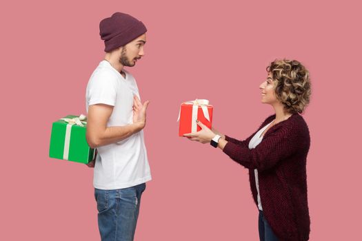 Side view portrait of amazing couple of friends in casual style standing, girl giving present box, boy hide behind gift, celebrate anniversary. Isolated, indoor, studio shot, pink background