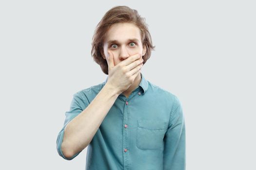 Portrait of shocked handsome long haired blonde young man in blue casual shirt standing, covering his mouth and looking at camera with big eyes. indoor studio shot, isolated on light grey background.