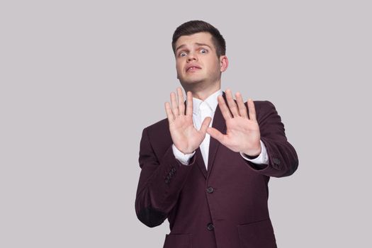 Please don't touch me. Portrait of scared handsome young man in suit and white shirt, standing, looking at camera with fear and blocking with hands. indoor studio shot, isolated on grey background.