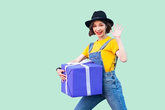 Cheerful happy young girl in hipster wear in denim overalls and black hat standing and trying to hold giant big heavy gift box and greeting you, showing five. Studio shot, green background, isolated