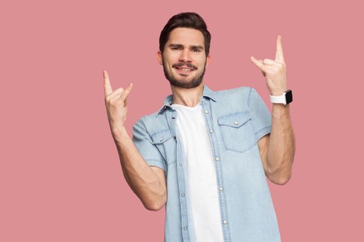 Portrait of happy handsome bearded young man in blue casual style shirt standing with rock sign and looking at camera with toothy smile. indoor studio shot, isolated on pink background.