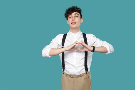 happy toothy smile man with heart hands sign gesture looking at camera. Portrait of handsome hipster curly young businessman in classic casual white shirt. studio shot isolated on blue background.