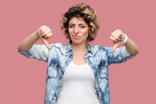 Dislike. Portrait of sad young woman with curly hairstyle in casual blue shirt standing, thumbs down and looking at camera with dissatisfied face. indoor studio shot, isolated on pink background.