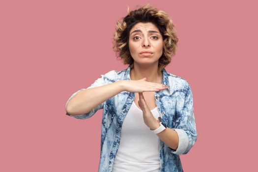 Timeout. I need more time. Portrait of worry pleased young woman with curly hairstyle in casual blue shirt standing with T gesture sign and looking at camera. studio shot, isolated on pink background