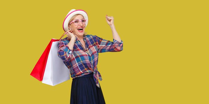 Make a great shopping! Satisfied modern granny in white hat and in checkered shirt holding shopping bags and triumphing with raised arm and toothy smile. Indoor, studio shot,yellow background,isolated