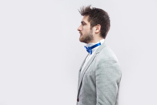 Profile side view portrait of serious handsome bearded man in casual grey suit, blue bow tie standing and looking straight wih calm serious face. indoor studio shot, isolated on light grey background.