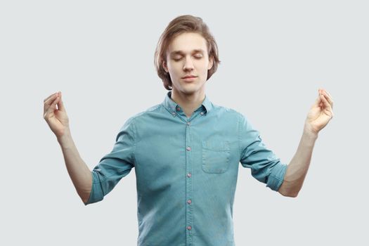 Portrait of calm handsome long haired blonde young man in blue casual shirt standing in yoga pose and meditating, and try to relax. indoor studio shot, isolated on light grey background.