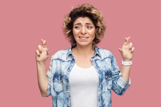 Portrait of hopeful young woman with curly hairstyle in casual blue shirt standing with crossed fingers, clenching teeth and looking away with worry face. studio shot, isolated on pink background.