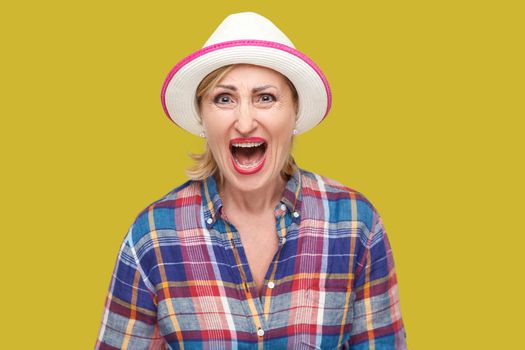 Portrait of angry or shocked modern stylish mature woman in casual style with white hat standing and looking at camera and screaming. indoor studio shot isolated on yellow background.