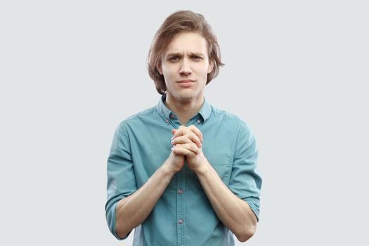 Please help me. Portrait of hopeful handsome long haired blonde young man in blue shirt standing with palm hand and hope for help or forgiveness. indoor studio shot, isolated on light grey background.