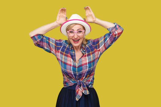 Portrait of funny modern stylish mature woman in casual style with hat and eyeglasses standing with bunny ears gesture and looking with toothy smile. indoor studio shot isolated on yellow background.