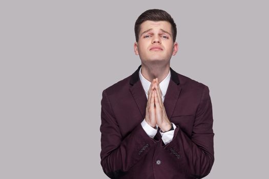 Portrait of pleasing worry handsome young man in violet suit and white shirt, standing, looking at camera with palms hand and pleading. indoor studio shot, isolated on grey background.