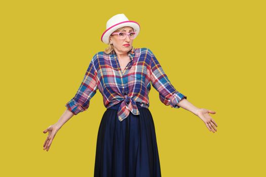 I don't know. Portrait of confused modern stylish mature woman in casual style with hat and eyeglasses standing, thinking, raised arms, looking away. indoor studio shot isolated on yellow background.