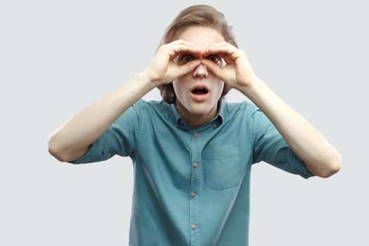 Portrait of amazed handsome long haired blonde young man in blue casual shirt standing with binoculars hands gesture with open mouth. indoor studio shot, isolated on light grey background.