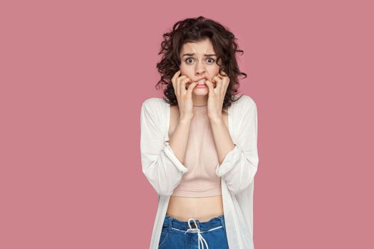 Portrait of nervous worried brunette young woman with curly hairstyle in casual style standing, confused face, bitting her nails and looking at camera. indoor studio shot isolated on pink background.