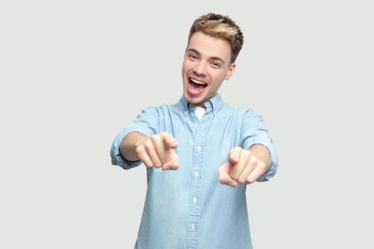 Portrait of surprised handsome young man in light blue shirt standing looking and pointing at camera with amazed happy face. indoor studio shot on grey background copy space.