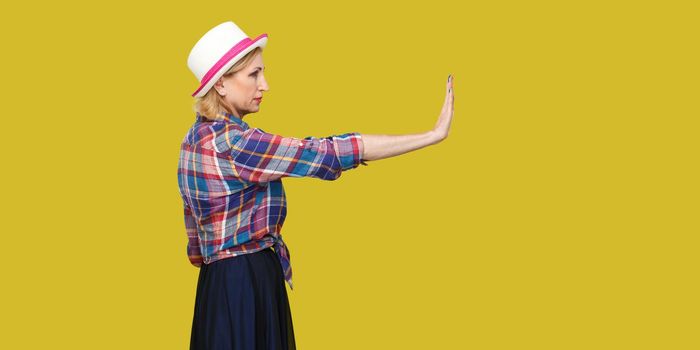 Stop it. Profile side view portrait of serious modern stylish mature woman in casual style with hat standing looking forward and showing stop sign. indoor studio shot isolated on yellow background.